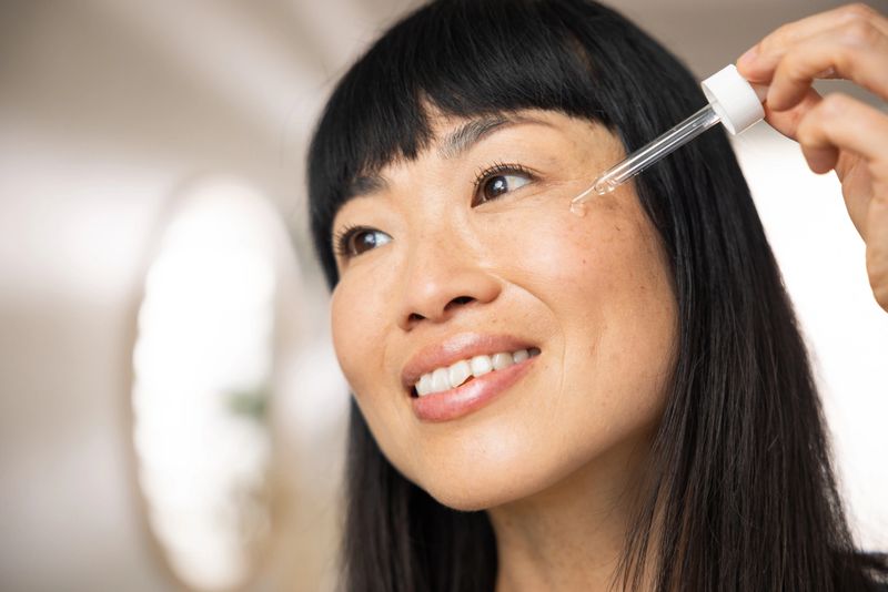 Asian woman applying serum on face in front of mirror in bathroom