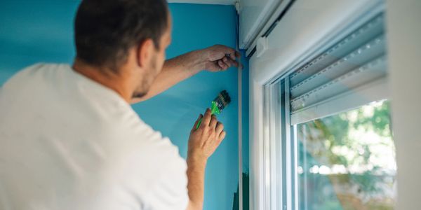 A man uses painter's tape to paint a room's corner near a window.