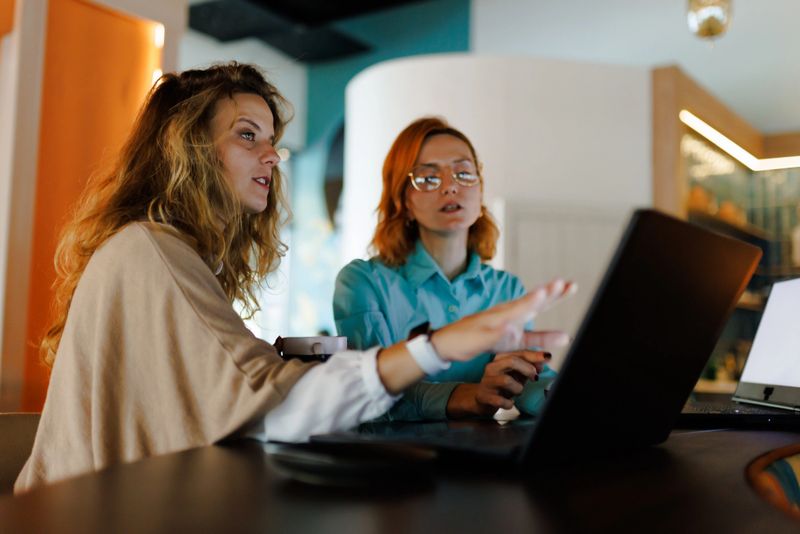 In a contemporary office setting, two women engage in a conversation, sharing ideas and strategies as they analyze data on a laptop. Their focused expressions convey professionalism and teamwork.