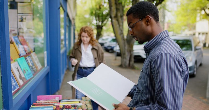 An African American man and a white woman interact with a small bookshelf outside of a bookstore in Oregon during the day
