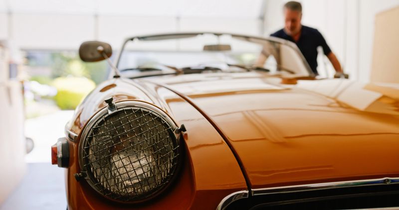 A low angle shot of a senior man taking a seat in his vintage convertible car inside his garage.