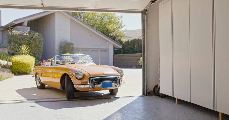 A wide shot of a vintage car backing out of a garage and driving away on a sunny day.