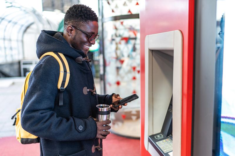 Young man using a smartphone to withdraw cash from an atm, holding a thermal mug filled with a hot beverage. Smiling and enjoying the convenience of modern banking in an urban setting
