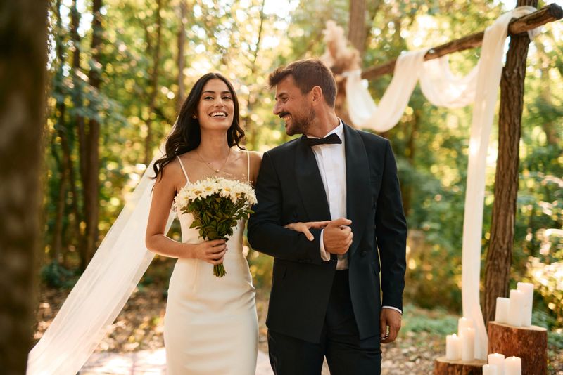 Smiling bride holding a vibrant bouquet, walking down the aisle beside her groom during a picturesque outdoor forest wedding ceremony, surrounded by nature's beauty and sunlight