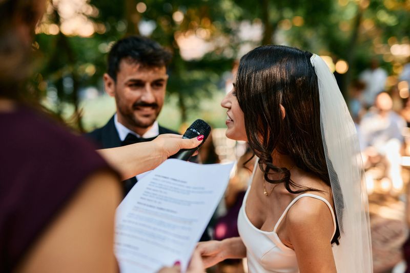 Young bride speaking into a microphone during her wedding ceremony, with the groom smiling in the background and the officiant holding the microphone and the wedding vows
