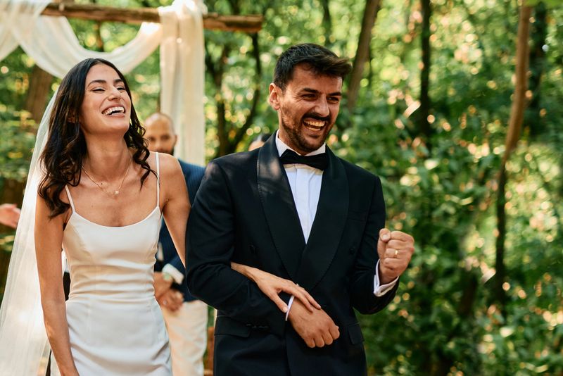 Happy newlywed couple laughing and walking down the aisle after their outdoor wedding ceremony in the woods, arm in arm and excited to start their new life together