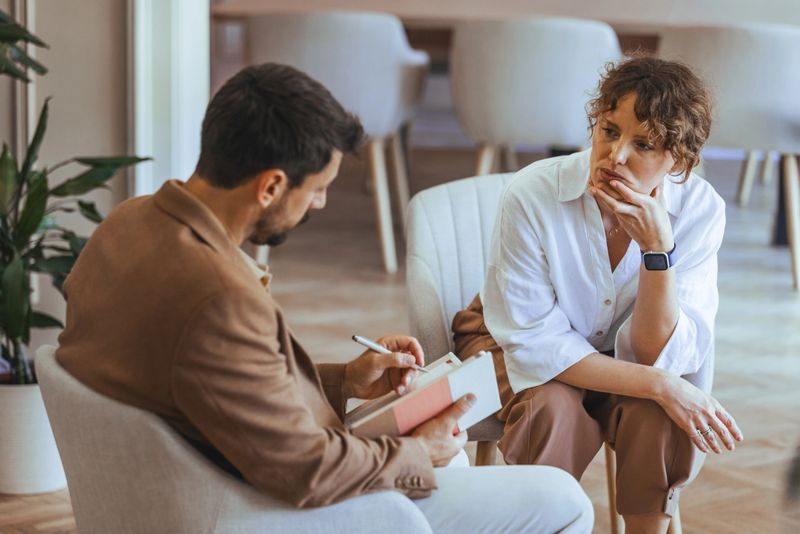 A counselor and a client are seated in a well-lit room, sharing a serious yet calm dialogue. The image portrays a professional setting focusing on therapy and support.