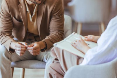 Man in brown jacket talking to a person with a notebook in a therapy session.