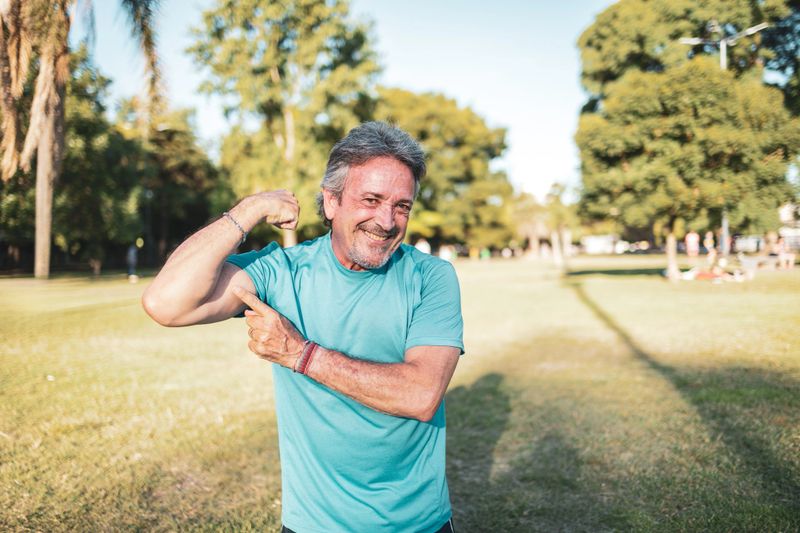 Happy mature man flexing his biceps and smiling joyfully at the camera while enjoying a sunny day in the park surrounded by nature