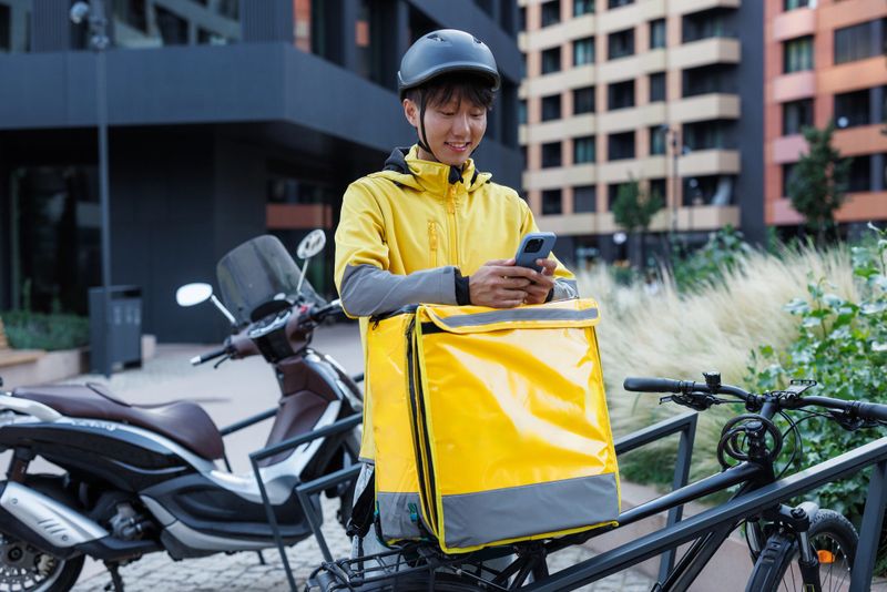 Courier using a smartphone while completing a delivery ride on a bicycle in a city. The scene highlights modern urban transportation solutions and the growing trend of food and goods delivery services.