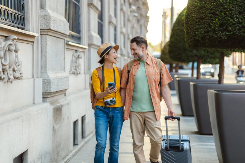 A cheerful couple strolling on an urban street together, embracing the spirit of travel and leisure on a sunny day. They carry luggage, radiating happiness and a sense of discovering new places.