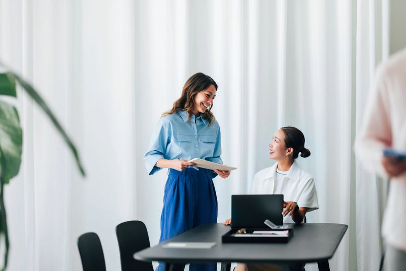 Two colleagues having a productive and enjoyable conversation around a desk in a contemporary office. The atmosphere depicts collaboration, positivity, and a shared sense of accomplishment in a professional environment.