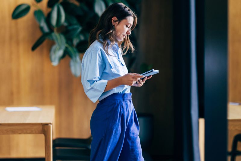 A woman dressed in professional attire stands indoors, focusing on her tablet in a modern office space with plants and warm wooden interior decor.