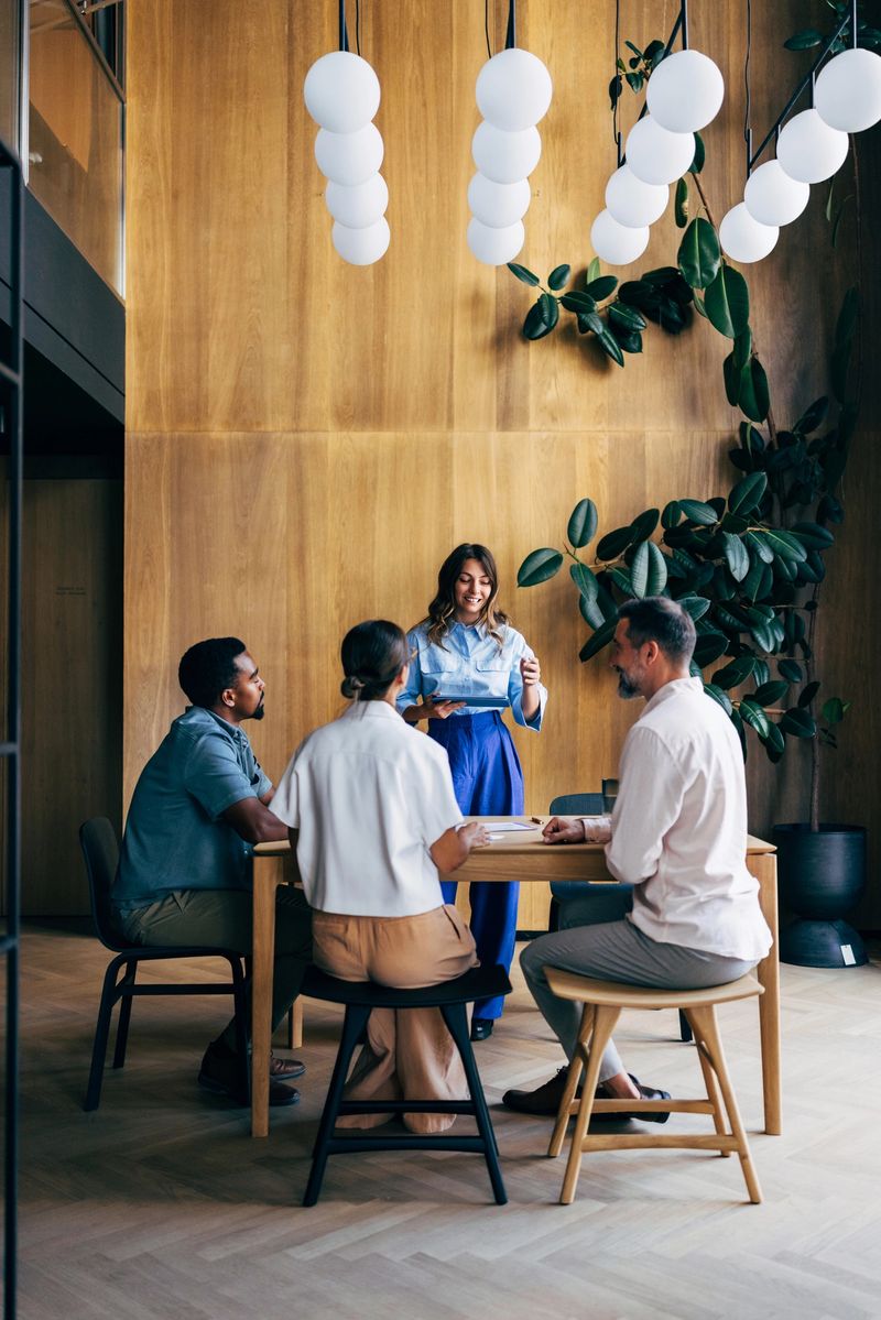 Professionals gather around a table in a modern office interior, brainstorming ideas beneath stylish lighting and surrounded by natural elements. The scene conveys teamwork, innovation, and a productive environment.