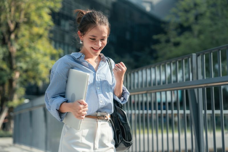 Confident young woman celebrating with joy as she reads the message on her smartphone saying that she's officially landed her first job. Overwhelmed with excitement, she throws her hands in the air, celebrating the beginning of a new chapter in her career