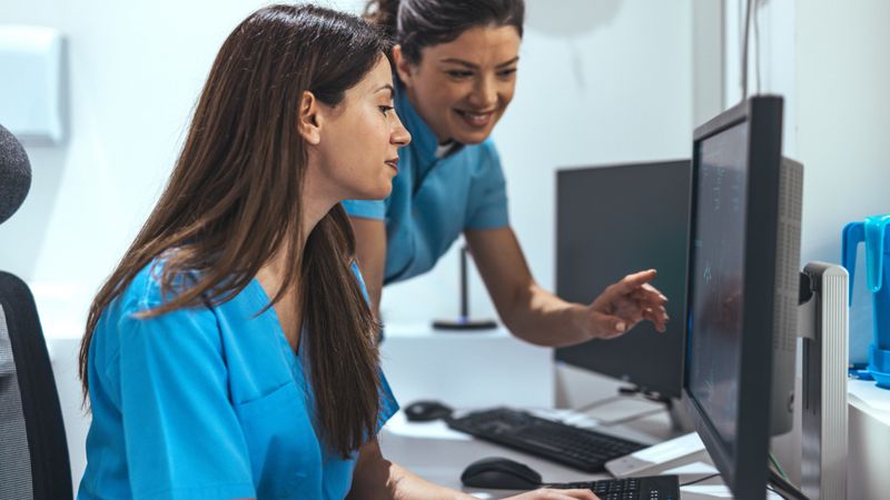 Two healthcare professionals in uniform reviewing data on a computer monitor in a clinical environment, displaying teamwork and collaboration essential in medical professions.