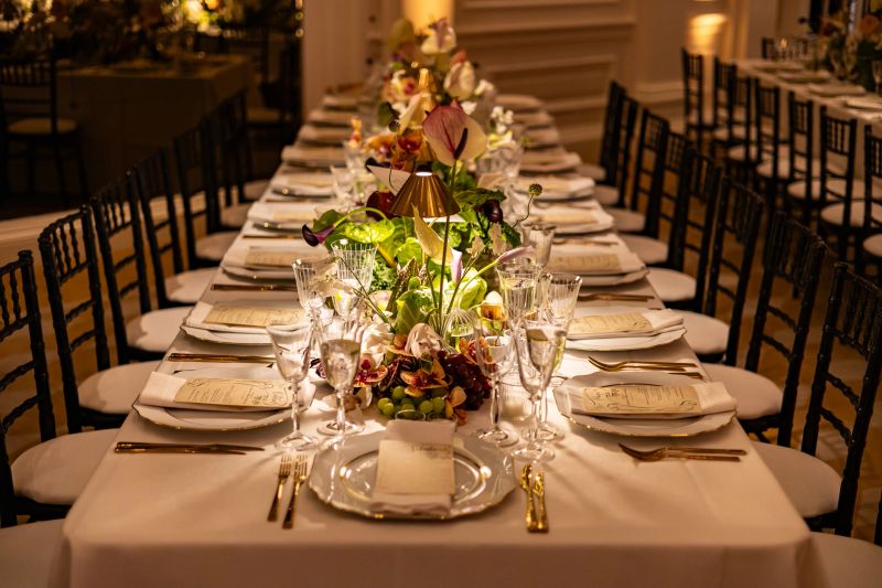 A beautifully arranged long dining table is set for a luxurious wedding dinner, featuring floral centerpieces, fine glassware, gold cutlery, and a romantic ambiance