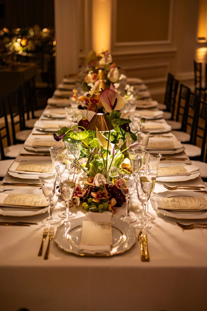 A beautifully arranged long dining table is set for a luxurious wedding dinner, featuring floral centerpieces, fine glassware, gold cutlery, and a romantic ambiance