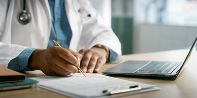 Doctor writing notes on a clipboard beside a laptop.