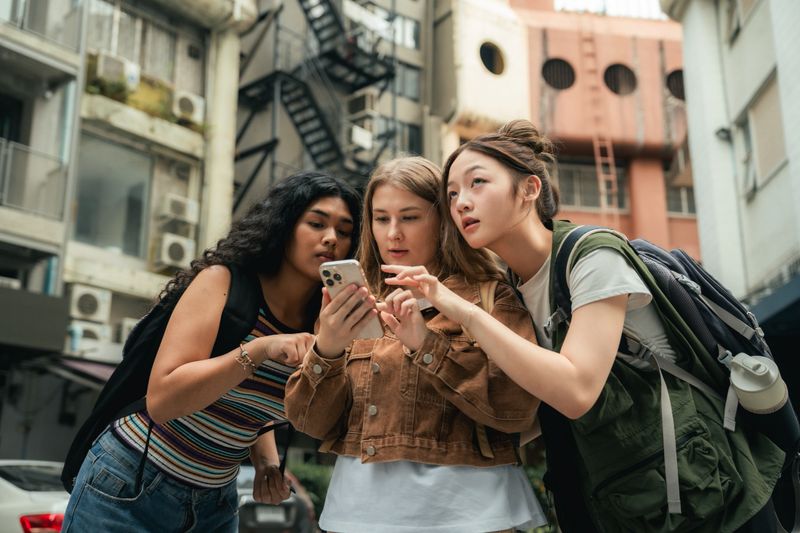 Group of friends gathered around a smartphone, looking at the map on the screen while navigating through the city, sharing laughs and a sense of adventure, relying on modern technology to guide their way while deepening their bond through the shared experience