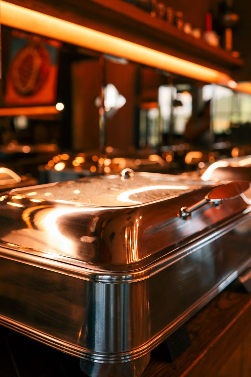 Close-up of stainless steel chafing dish at buffet in restaurant. Warm lighting reflects on metal surface, creating cozy dining atmosphere with catering equipment and utensils in background.