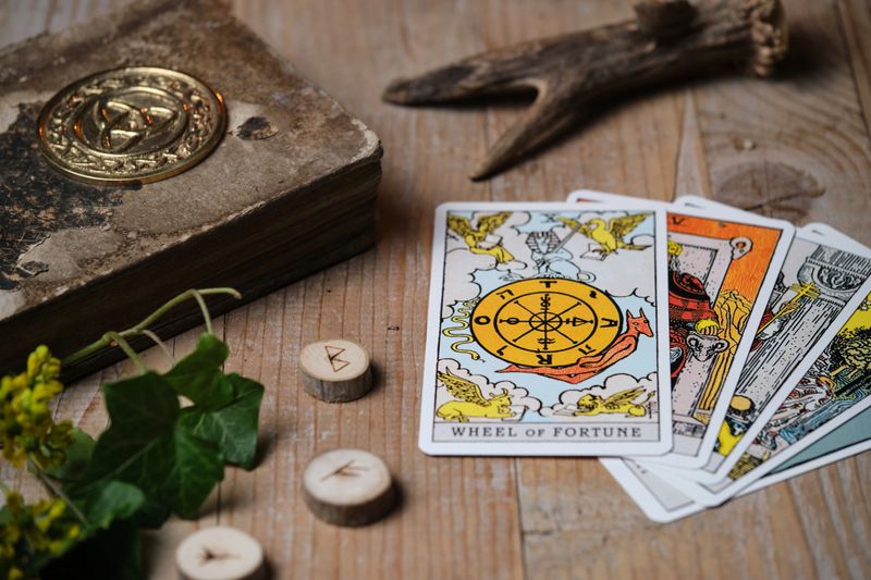 A moody, atmospheric scene featuring tarot cards laid out on an old wooden table, with the "Wheel of Fortune" card placed on top. Surrounded by rune stones and an ancient book embossed with a Celtic triquetra symbol. Warm lighting and dark, mystical tones create a spiritual and magical ambiance. Perfect for themes like divination, mysticism, witchcraft, or the occult.