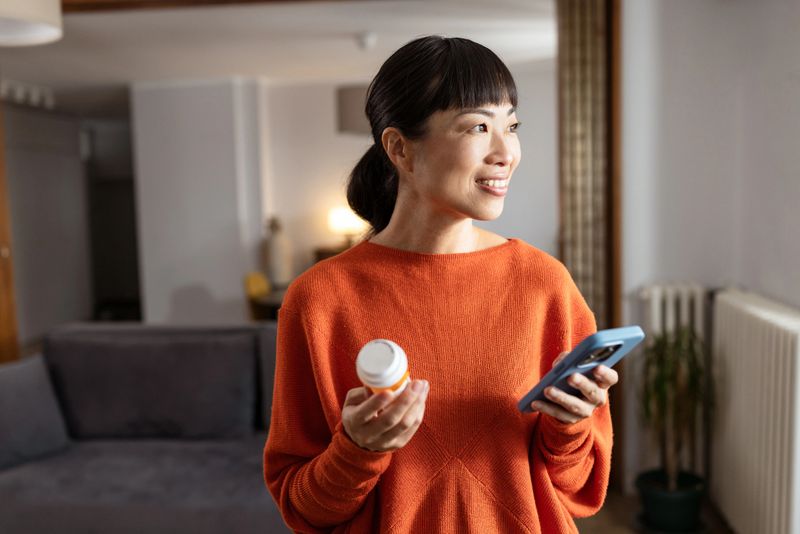 A cheerful Asian woman wearing an orange sweater, holding a smartphone and a pill bottle. She is standing in a stylish living room, appearing thoughtful and happy while looking towards the window.