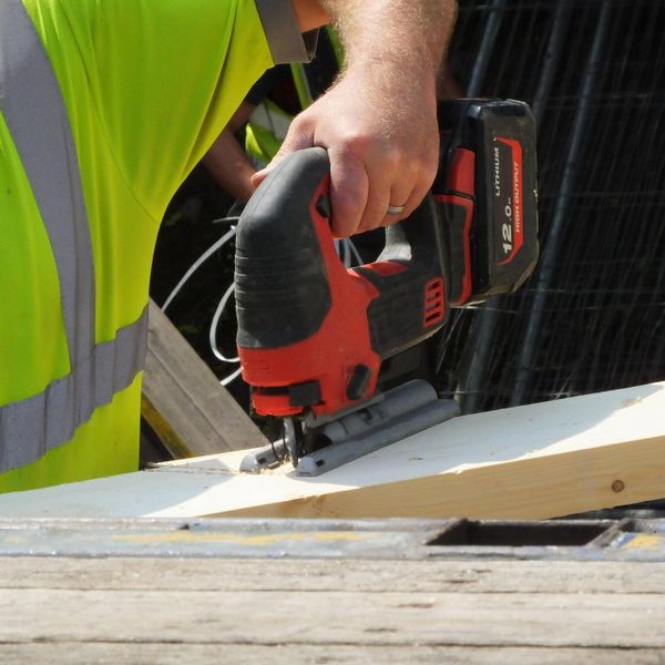 Worker using a red and black cordless jigsaw on wood.