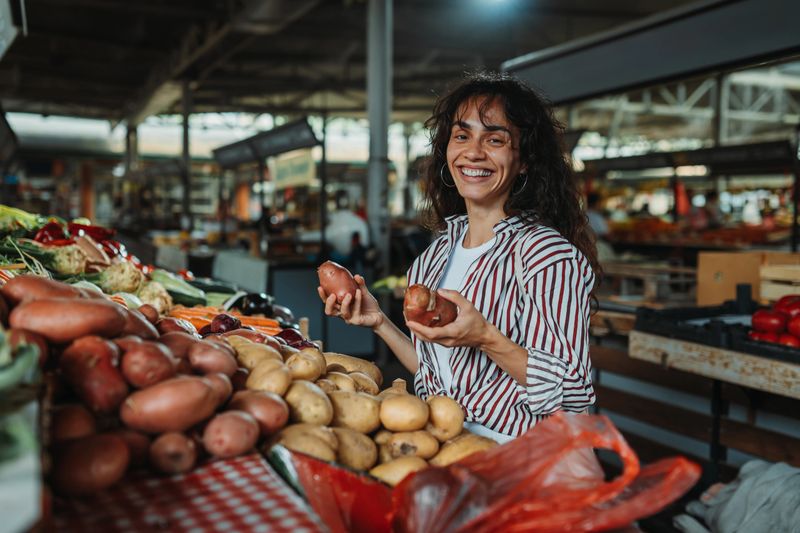 Happy woman buying potatoes at the farmers market, holding two potatoes in her hands, smiling at camera