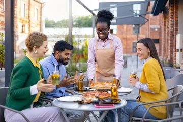 A waitress serves pizza to happy friends dining outdoors at a table.