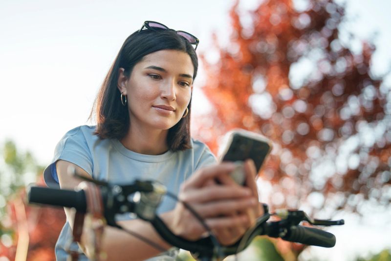 A young woman pauses in a public park, standing still on her bicycle while using her smartphone. The scene blends urban leisure with digital connection in a natural setting.