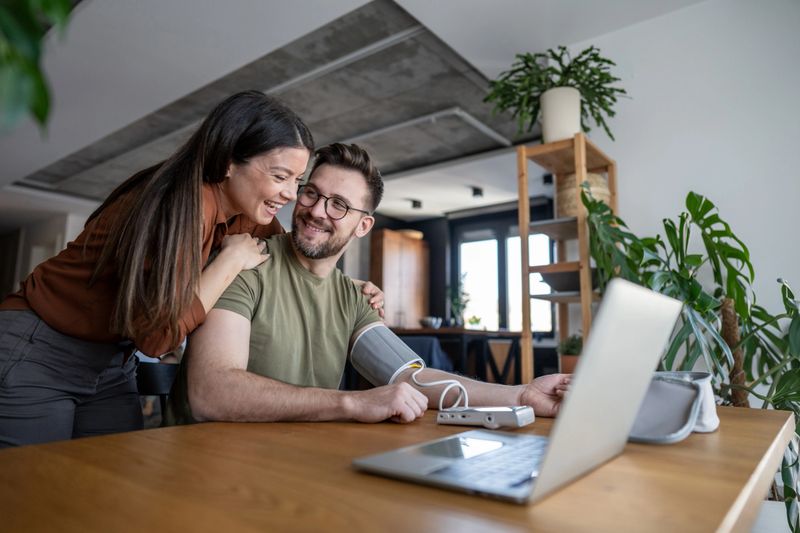 Smiling young couple is using a digital sphygmomanometer and a laptop for an online medical consultation with their doctor, checking blood pressure from home
