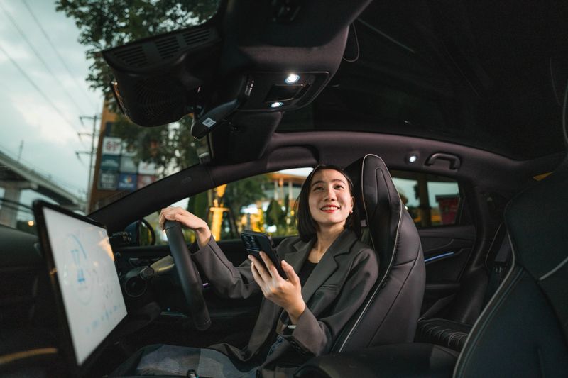 A woman sitting in her EV car, exploring various features on the car's smart application through her smartphone, including navigation, climate control, and energy-saving options