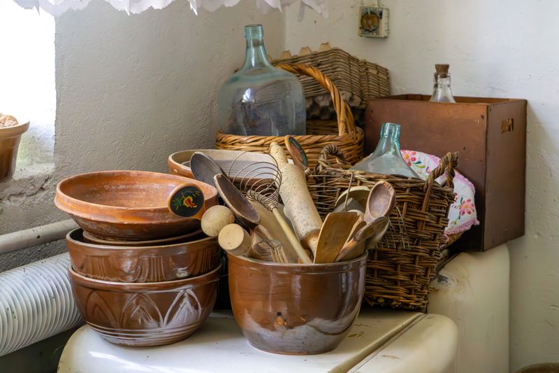 Rustic kitchen set on a windowsill – clay bowls, wooden spoons, and glass bottles in wicker baskets, arranged against a whitewashed wall in a countryside style