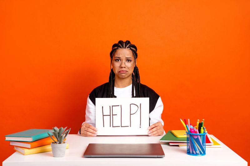 A young girl sitting at her desk, holding a sign reflecting her need for help, surrounded by colorful school supplies.