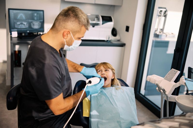 A young girl with blonde hair is seated in a modern dental chair, receiving 3d scanning from a male dentist wearing scrubs and gloves in a clean clinic.