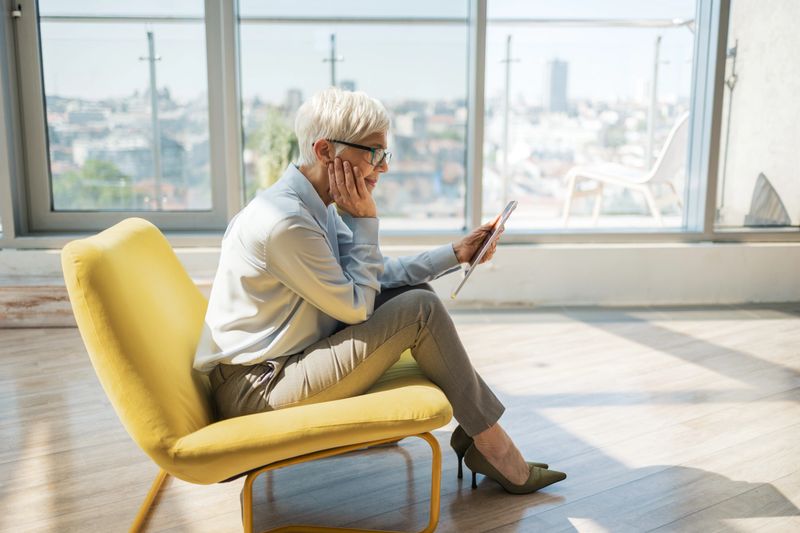 A professional woman is seated on a modern yellow chair, reading from a tablet in a contemporary office with large windows offering a panoramic urban view in the background.