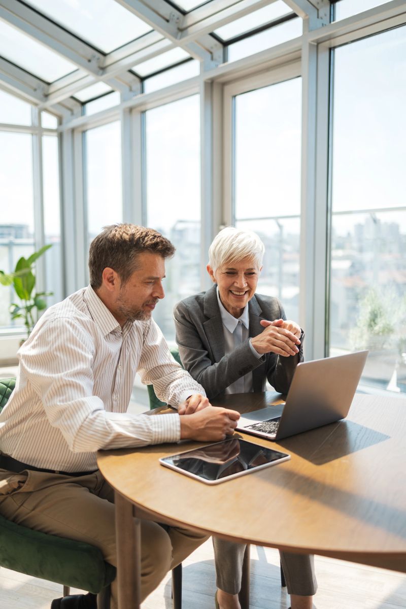 Two business professionals seated in a bright office, engaging in teamwork and collaboration, with modern technology and a laptop emphasizing productivity and cooperation.