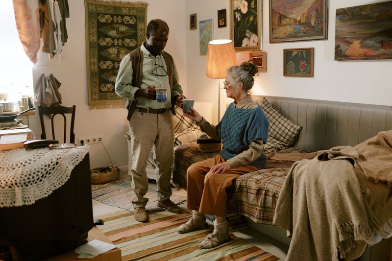 Senior Black man standing and holding mug, while senior Caucasian woman sitting on bed and reaching out with cup in modest living space decorated with paintings and textiles