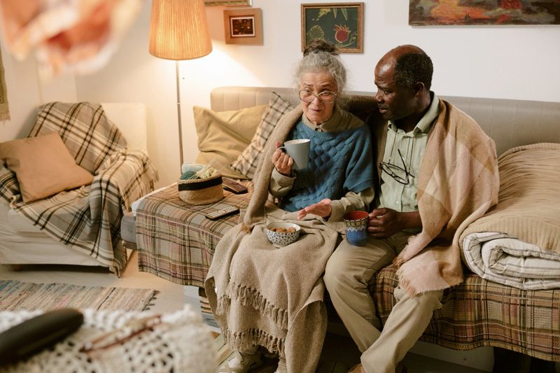 Senior Caucasian woman and senior Black man sitting together on sofa wrapped in blankets, holding mugs, sharing quiet moment in modest living room, looking slightly off camera