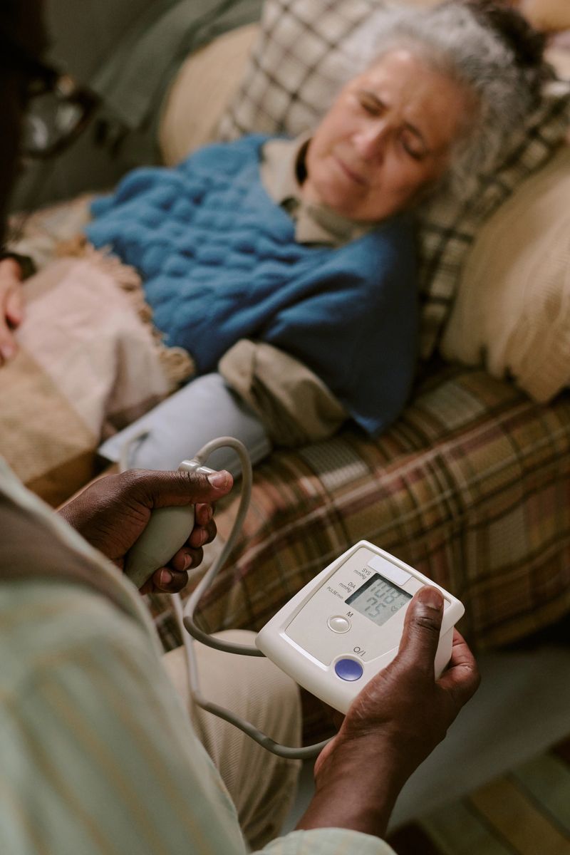 Senior Caucasian woman lying in bed receiving care from Black man measuring blood pressure with digital monitor, elderly patient resting while caregiver checking health status