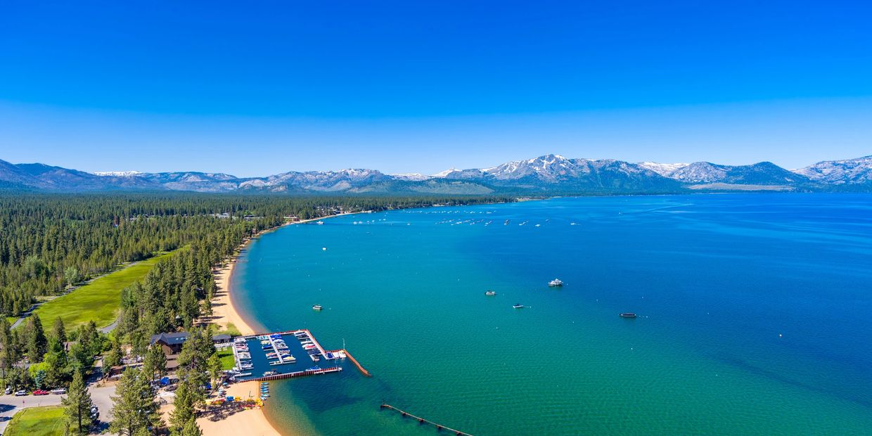 A stunning lake with boats, surrounded by pine forests and snow-capped mountains under a clear blue sky.
