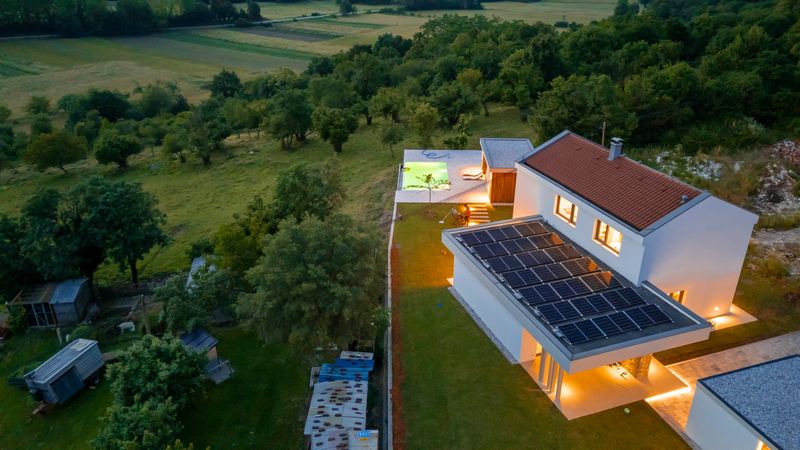 An eco-friendly contemporary house with solar panels, illuminated by warm lighting during blue hour, surrounded by lush greenery and a peaceful rural landscape.