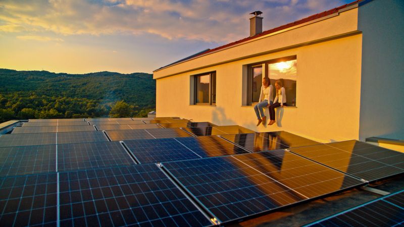 Panoramic view of mid-aged woman and young daughter sitting on window ledge overlooking residential solar panel installation, bathed in warm sunset light against mountainous backdrop.