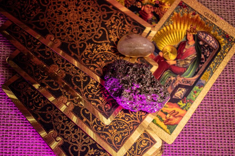 Tarot cards with gold designs of a King and Queen lie on woven fabric. Rock crystal, pyrite, and a lit candle glow in purple light, evoking a mystical, spiritual ritual scene.