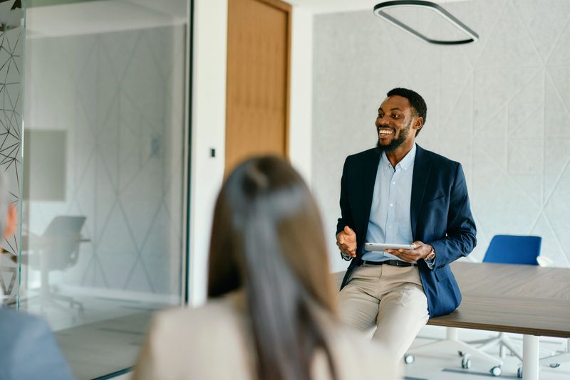Young businessman holding a tablet, leading a meeting with colleagues in a modern office. Sitting on the desk, smiling and fostering a positive atmosphere for collaboration and innovation
