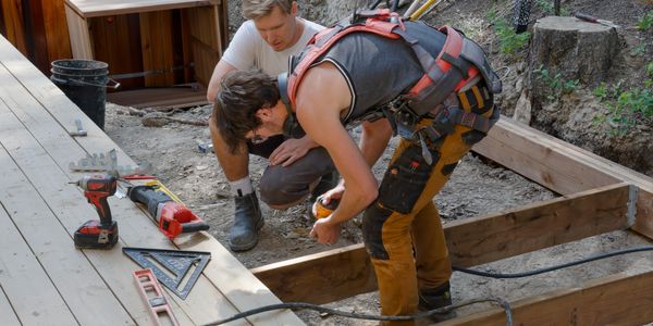 Two construction workers measuring wood for a deck frame outdoors.