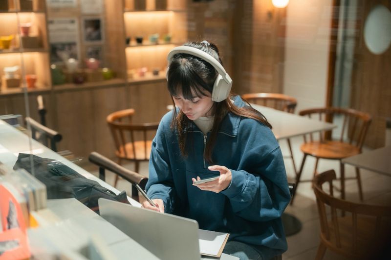 Gen Z woman multitasks in a co-working space with quiet focus and creative energy, listening to an audiobook through wireless headphones, taking notes in her book, and studying with her laptop, blending self-study, digital learning, and aesthetic productivity