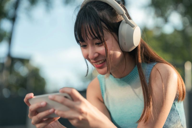 Cheerful Gen Z woman plays a mobile game on her smartphone outdoors, enjoying leisure time, digital entertainment, and casual gaming during a break, reflecting mobile lifestyle, screen time habits, and gaming culture