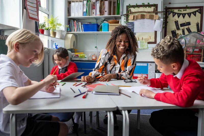 A wide shot of a female teacher sitting down with her students as they work in their textbooks during a lesson. The teacher is wearing smart-casual clothing and has brown afro-style hair. The students are wearing their school uniform, which consists of a red jumper and a white shirt.Videos are available similar to this scenario.